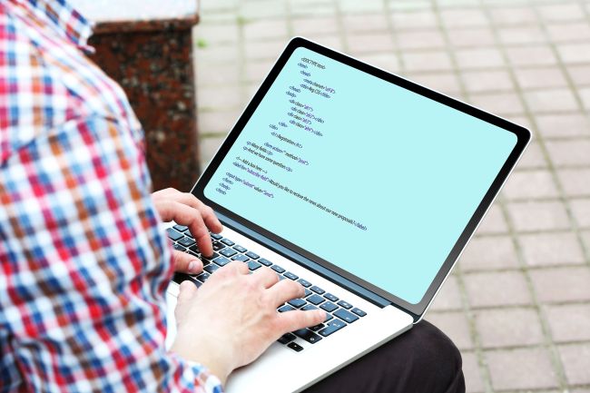 Person in a red and blue plaid button down shirt works on their laptop while sitting outside on a bench.