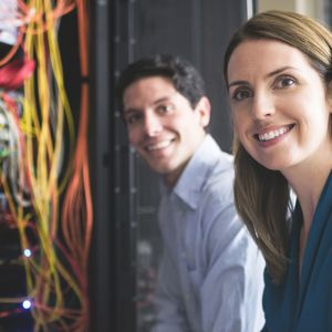 CompTIA A+ with ITIL Foundation image of two IT professionals female and male smiling with colorful lights and cords hanging from wall of hard drives